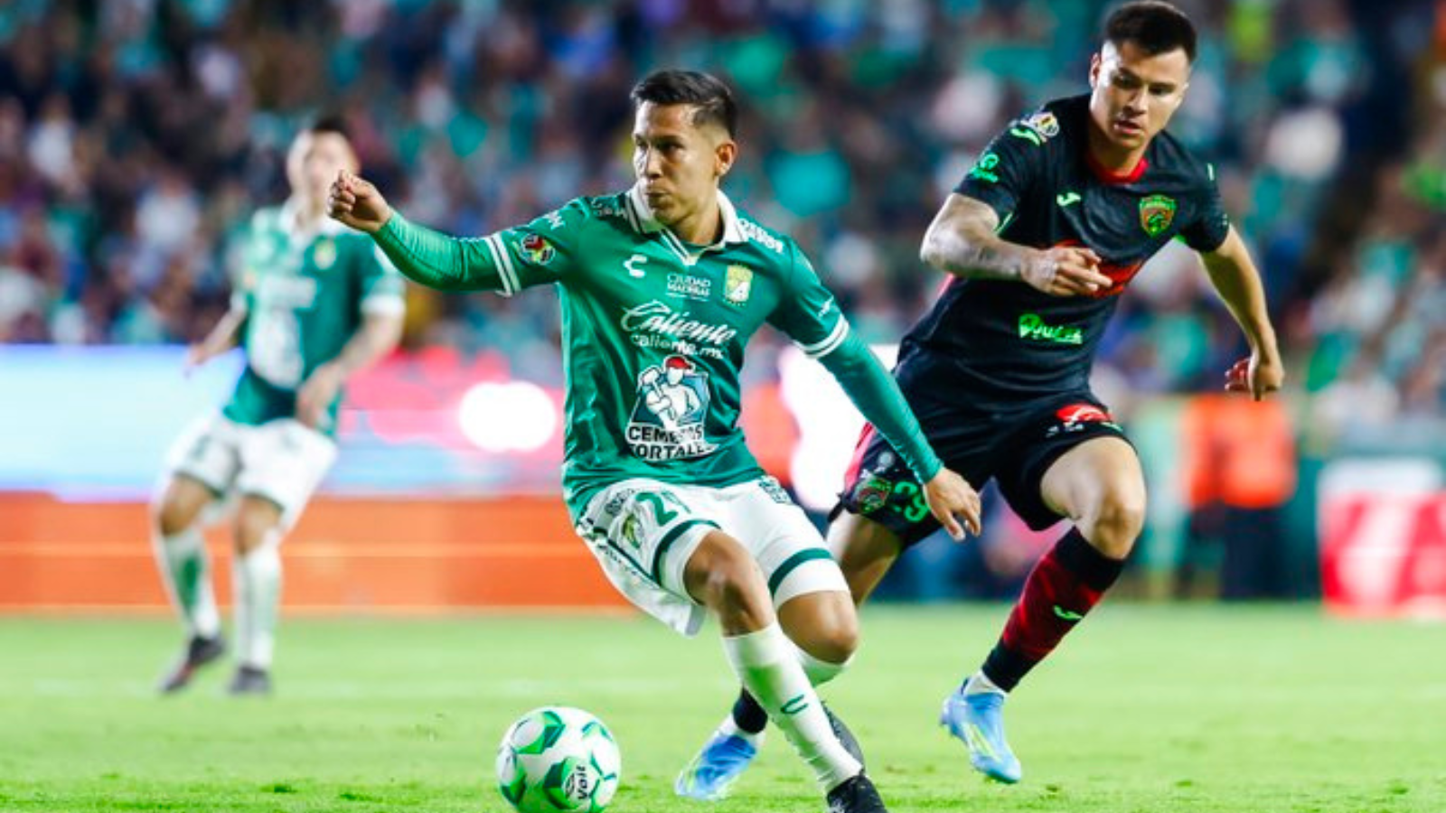 Two soccer players chase a green-and-white ball on the field during a match, with a blurred crowd in the background.