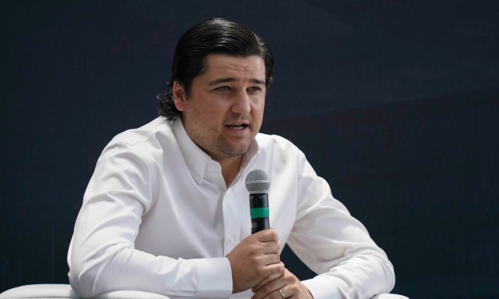 Man in a white button-down shirt speaks into a handheld microphone against a dark background.