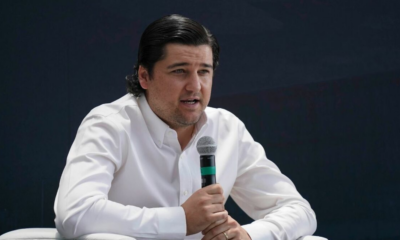 Man in a white button-down shirt speaks into a handheld microphone against a dark background.