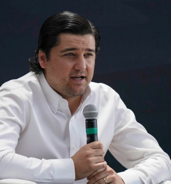 Man in a white button-down shirt speaks into a handheld microphone against a dark background.