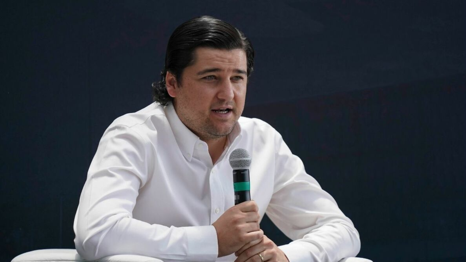 Man in a white button-down shirt speaks into a handheld microphone against a dark background.