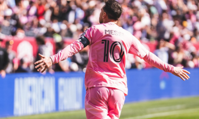 Messi in a pink jersey with number 10, arms outstretched, celebrating on a soccer field with a cheering crowd in the background