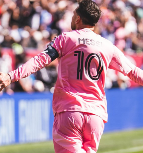 Messi in a pink jersey with number 10, arms outstretched, celebrating on a soccer field with a cheering crowd in the background