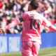 Messi in a pink jersey with number 10, arms outstretched, celebrating on a soccer field with a cheering crowd in the background