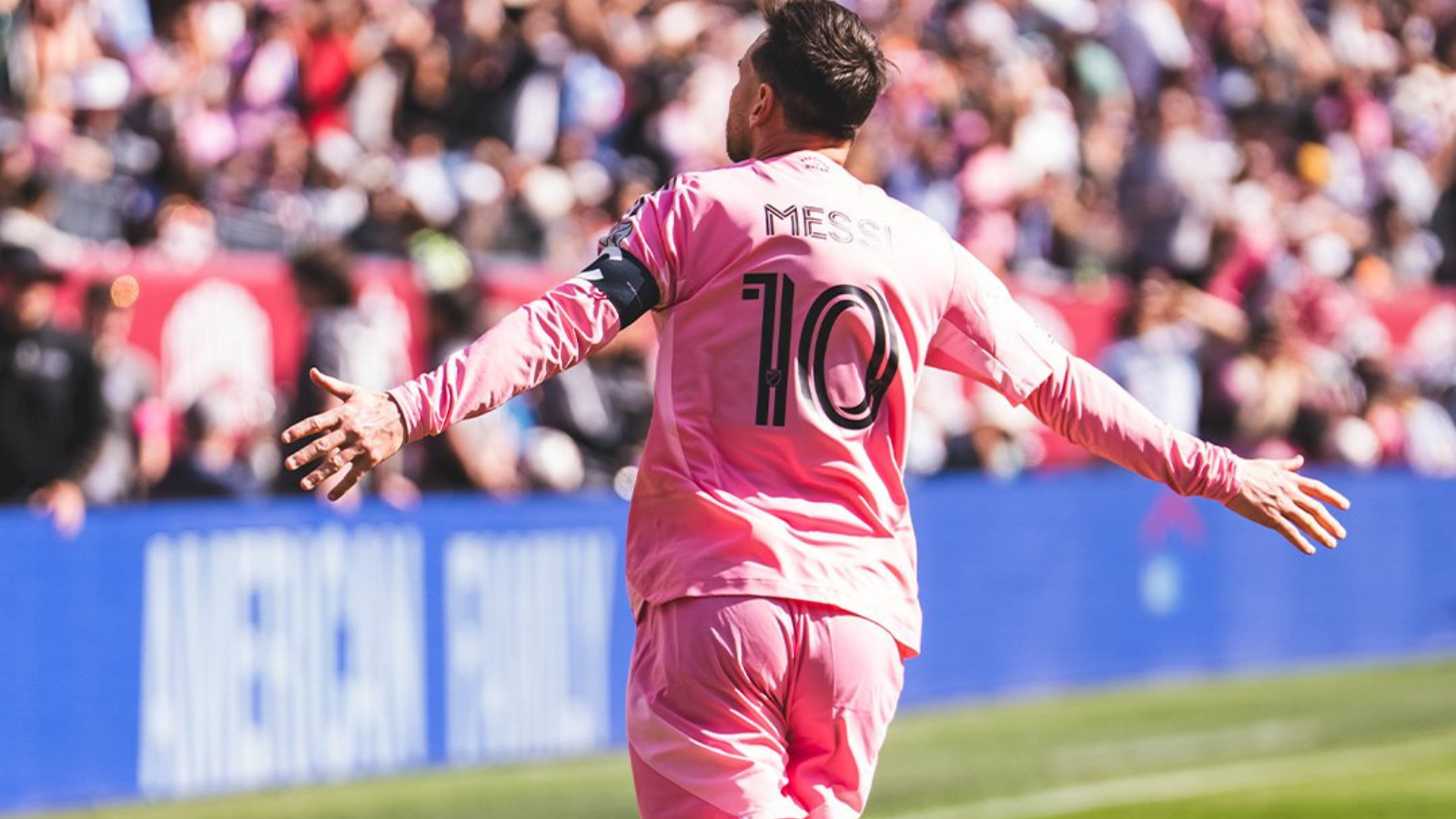 Messi in a pink jersey with number 10, arms outstretched, celebrating on a soccer field with a cheering crowd in the background