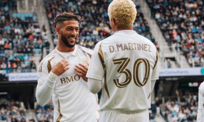 Smiling soccer players in white and gold kits chat on the field with a large crowd in the stands behind them.