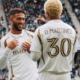 Smiling soccer players in white and gold kits chat on the field with a large crowd in the stands behind them.