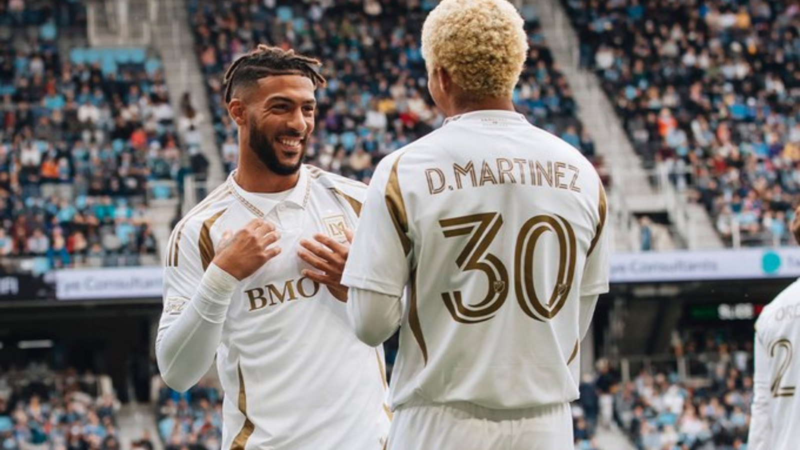 Smiling soccer players in white and gold kits chat on the field with a large crowd in the stands behind them.