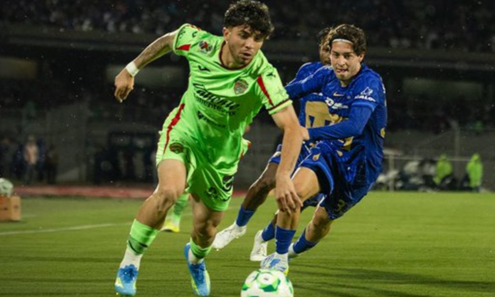 Two soccer players chase a ball on a green field during a night match; one in neon green kit, one in blue, in a close contest.