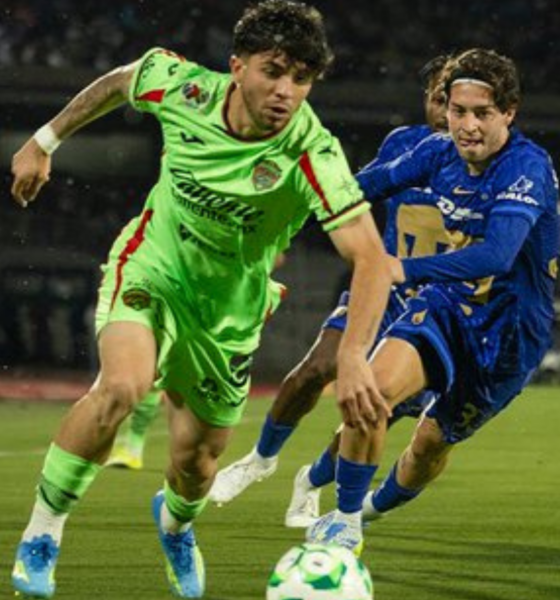 Two soccer players chase a ball on a green field during a night match; one in neon green kit, one in blue, in a close contest.