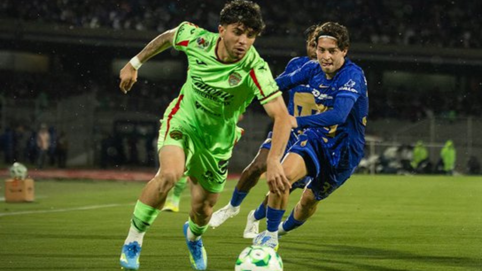 Two soccer players chase a ball on a green field during a night match; one in neon green kit, one in blue, in a close contest.