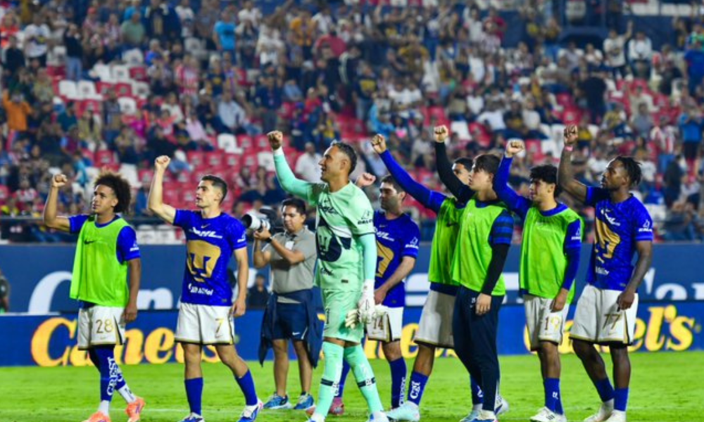 Soccer players in blue jerseys celebrate on the field with raised fists as a cheering crowd watches from the stands.