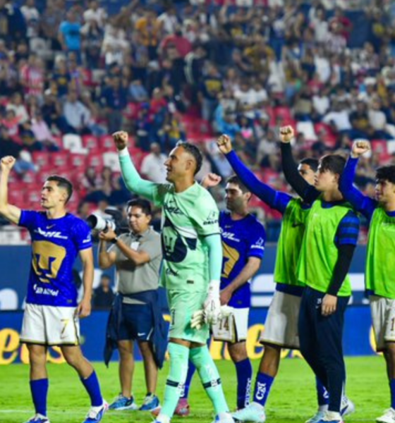 Soccer players in blue jerseys celebrate on the field with raised fists as a cheering crowd watches from the stands.
