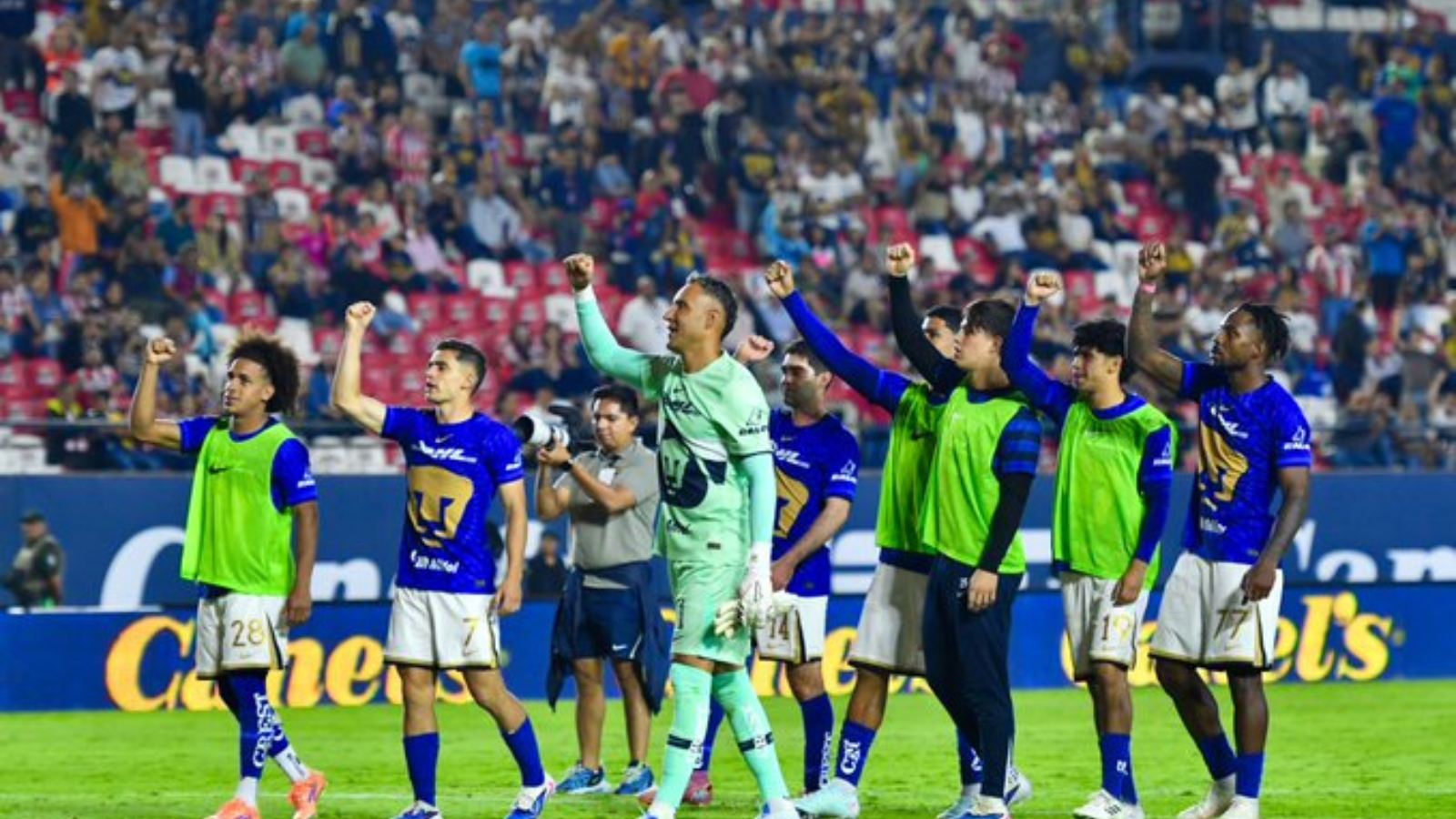 Soccer players in blue jerseys celebrate on the field with raised fists as a cheering crowd watches from the stands.