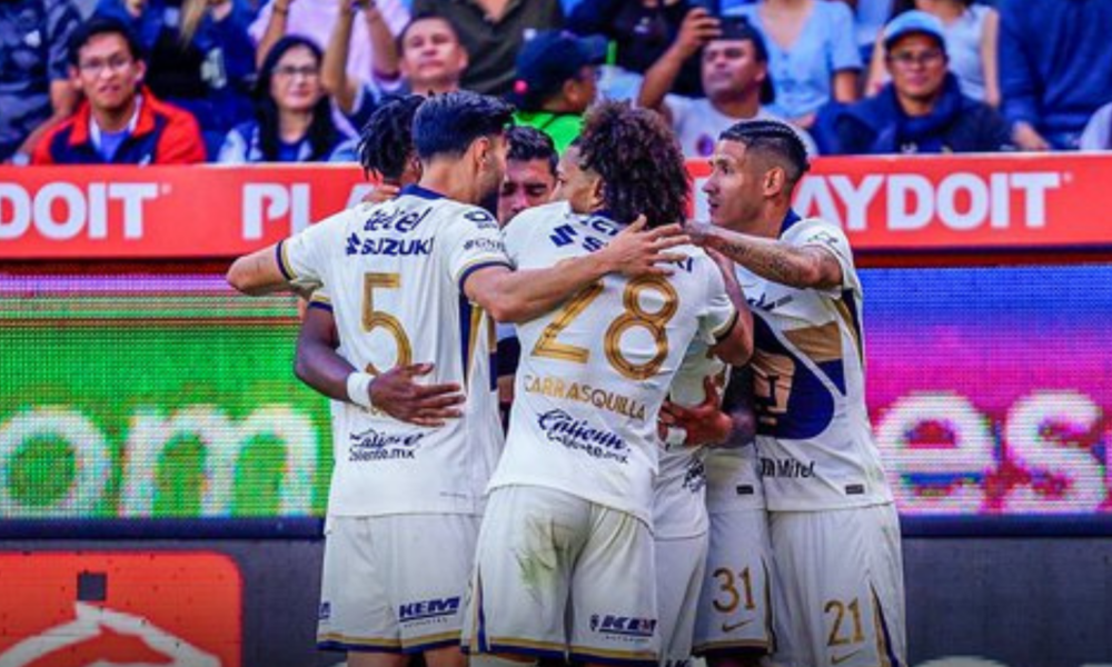 Soccer teammates in white uniforms hug and celebrate on the field amid a crowd and bright advertising boards behind them.