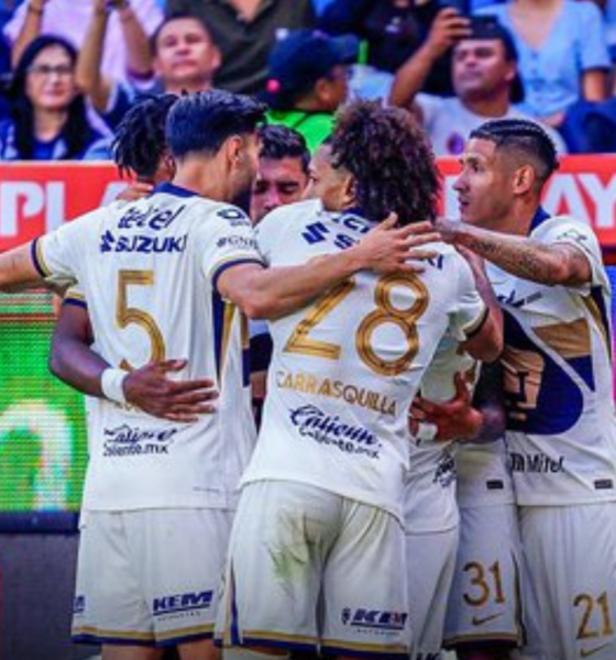 Soccer teammates in white uniforms hug and celebrate on the field amid a crowd and bright advertising boards behind them.
