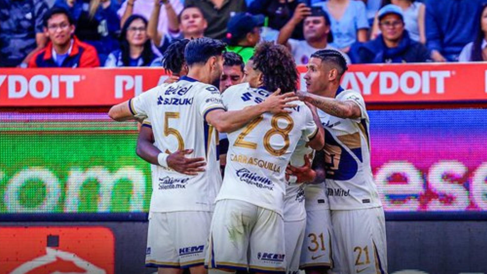 Soccer teammates in white uniforms hug and celebrate on the field amid a crowd and bright advertising boards behind them.