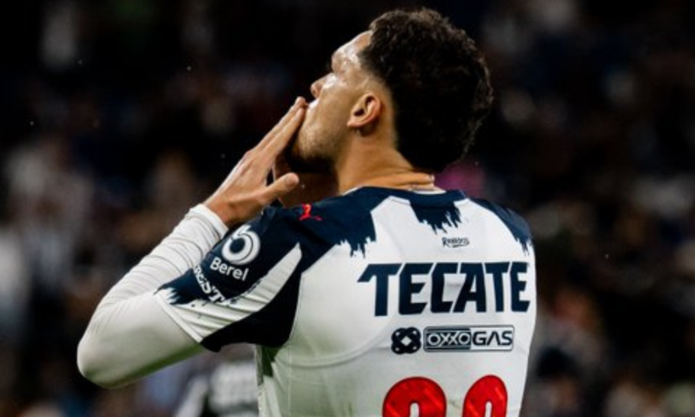 Back view of a soccer player in a white and navy Tecate jersey, hands near his face in a stadium celebration.