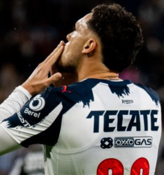 Back view of a soccer player in a white and navy Tecate jersey, hands near his face in a stadium celebration.