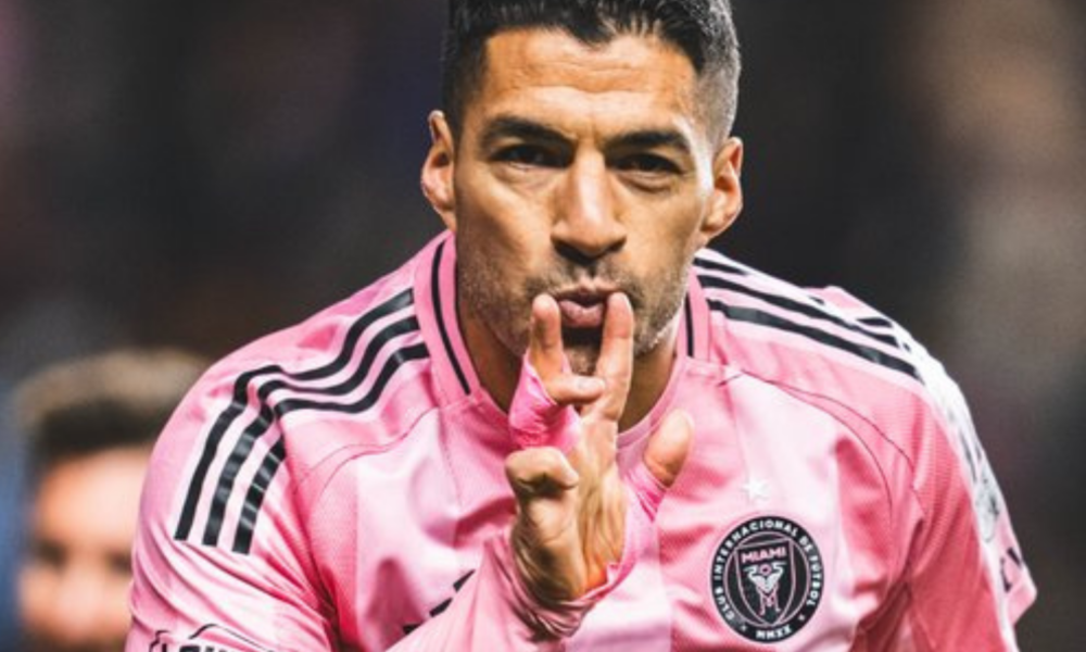 Soccer player in a pink jersey celebrates on the field, making a kiss gesture with his fingers closer to his lips.