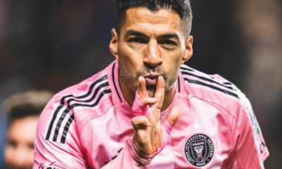 Soccer player in a pink jersey celebrates on the field, making a kiss gesture with his fingers closer to his lips.