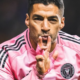 Soccer player in a pink jersey celebrates on the field, making a kiss gesture with his fingers closer to his lips.
