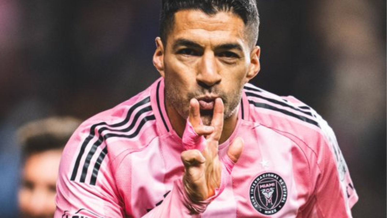 Soccer player in a pink jersey celebrates on the field, making a kiss gesture with his fingers closer to his lips.