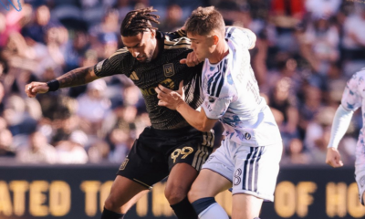 Two male soccer players clash for the ball in a crowded stadium; one in a black kit with gold accents, the other in white/blue stripes.