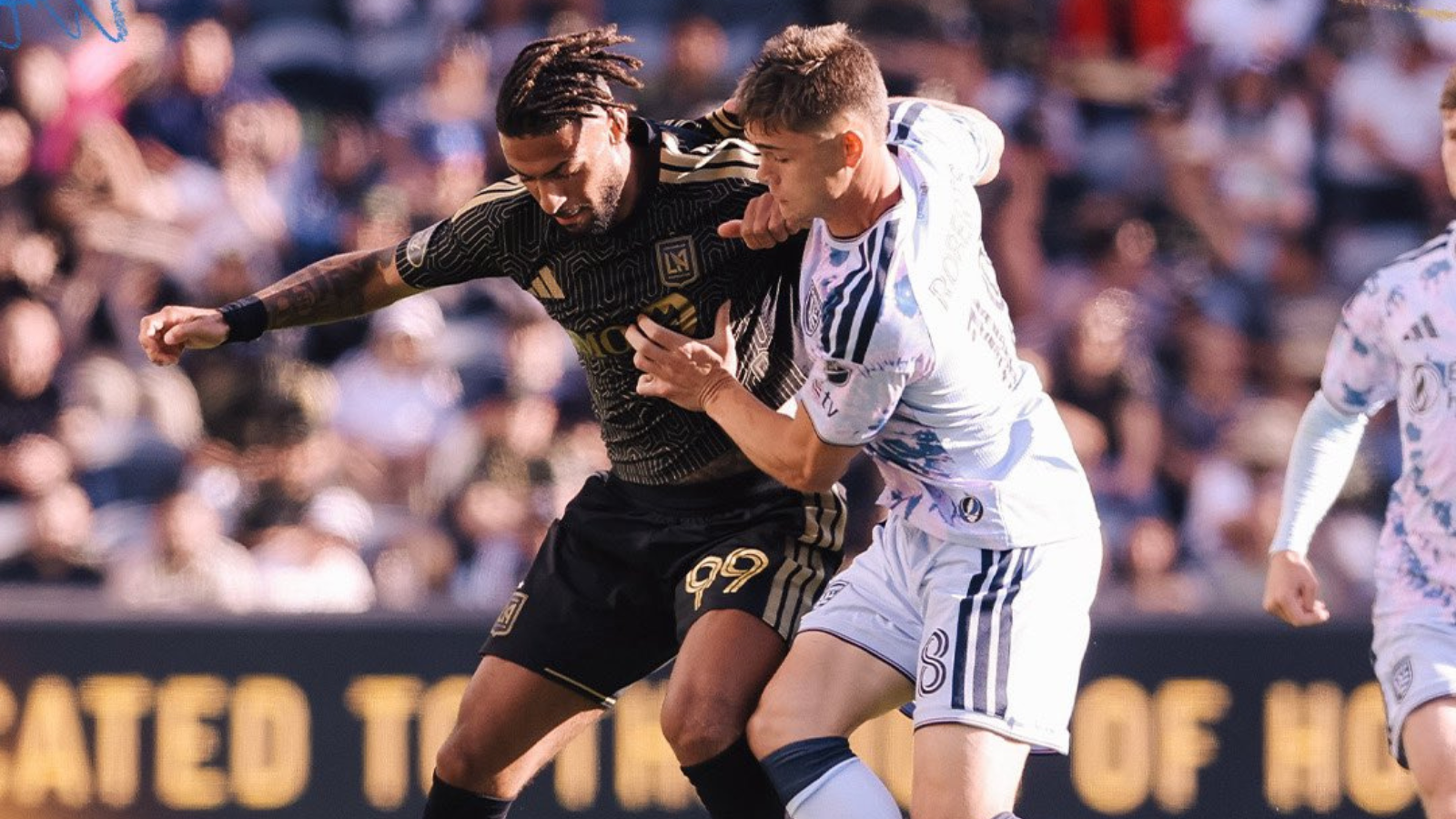 Two male soccer players clash for the ball in a crowded stadium; one in a black kit with gold accents, the other in white/blue stripes.