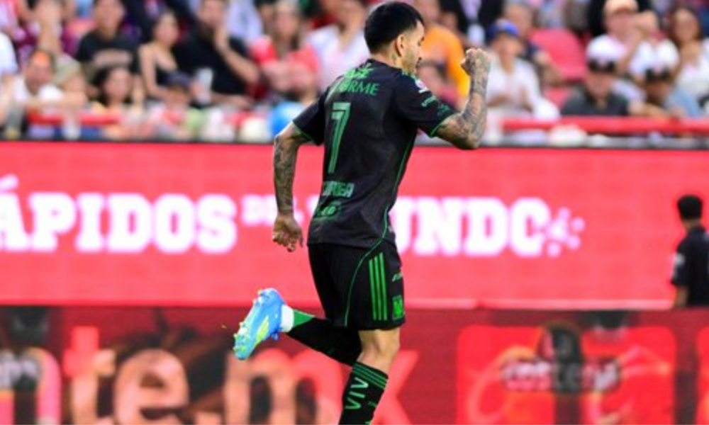 Soccer player in a black kit with green trim (#7) celebrating a goal, running on the field with blurred fans in the stands behind him.