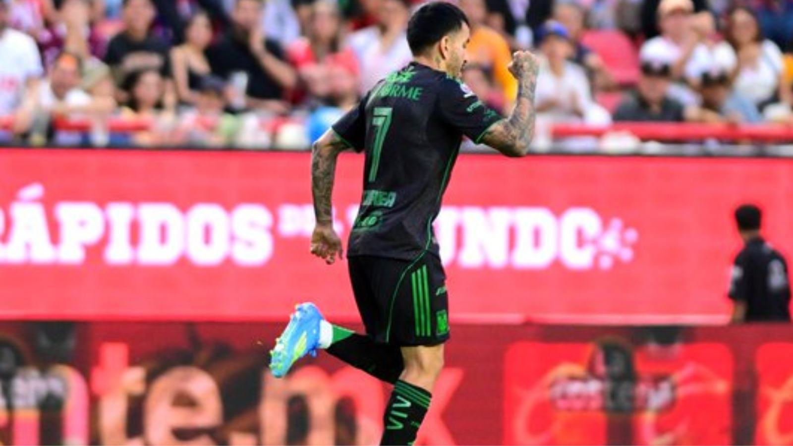 Soccer player in a black kit with green trim (#7) celebrating a goal, running on the field with blurred fans in the stands behind him.