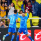 Two soccer players in blue jerseys celebrate a goal while high-fiving on the pitch as cheering fans look on from the stands behind a barrier.