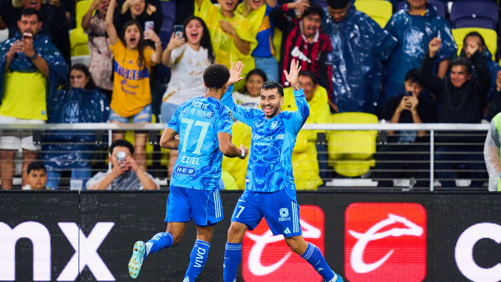 Two soccer players in blue jerseys celebrate a goal while high-fiving on the pitch as cheering fans look on from the stands behind a barrier.