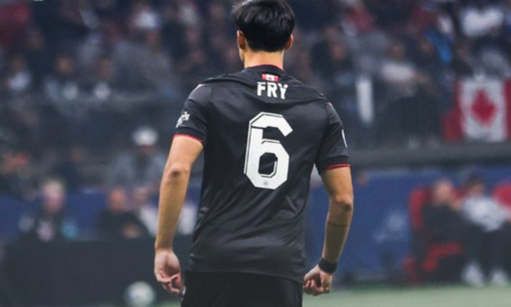 Soccer player seen from behind in a black jersey with the white number 6 and the name 'FRY' on the back, on a blurred stadium background.