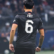 Soccer player seen from behind in a black jersey with the white number 6 and the name 'FRY' on the back, on a blurred stadium background.