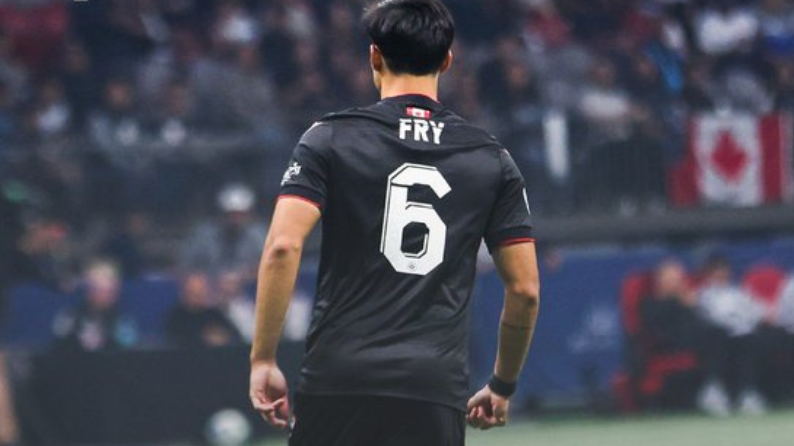 Soccer player seen from behind in a black jersey with the white number 6 and the name 'FRY' on the back, on a blurred stadium background.