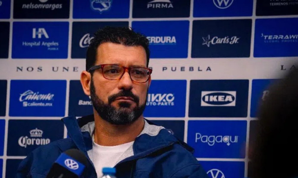 Man with red-framed glasses speaks at a press conference in front of a blue sponsor banner backdrop.