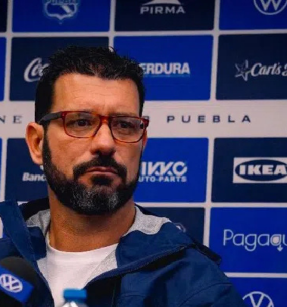 Man with red-framed glasses speaks at a press conference in front of a blue sponsor banner backdrop.