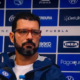 Man with red-framed glasses speaks at a press conference in front of a blue sponsor banner backdrop.