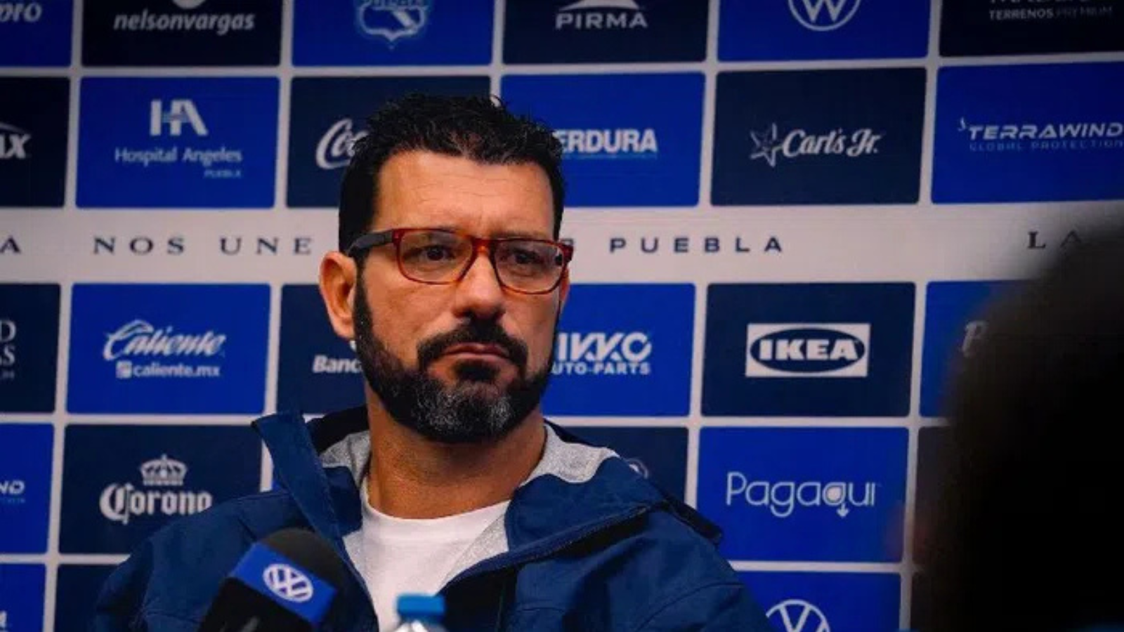 Man with red-framed glasses speaks at a press conference in front of a blue sponsor banner backdrop.