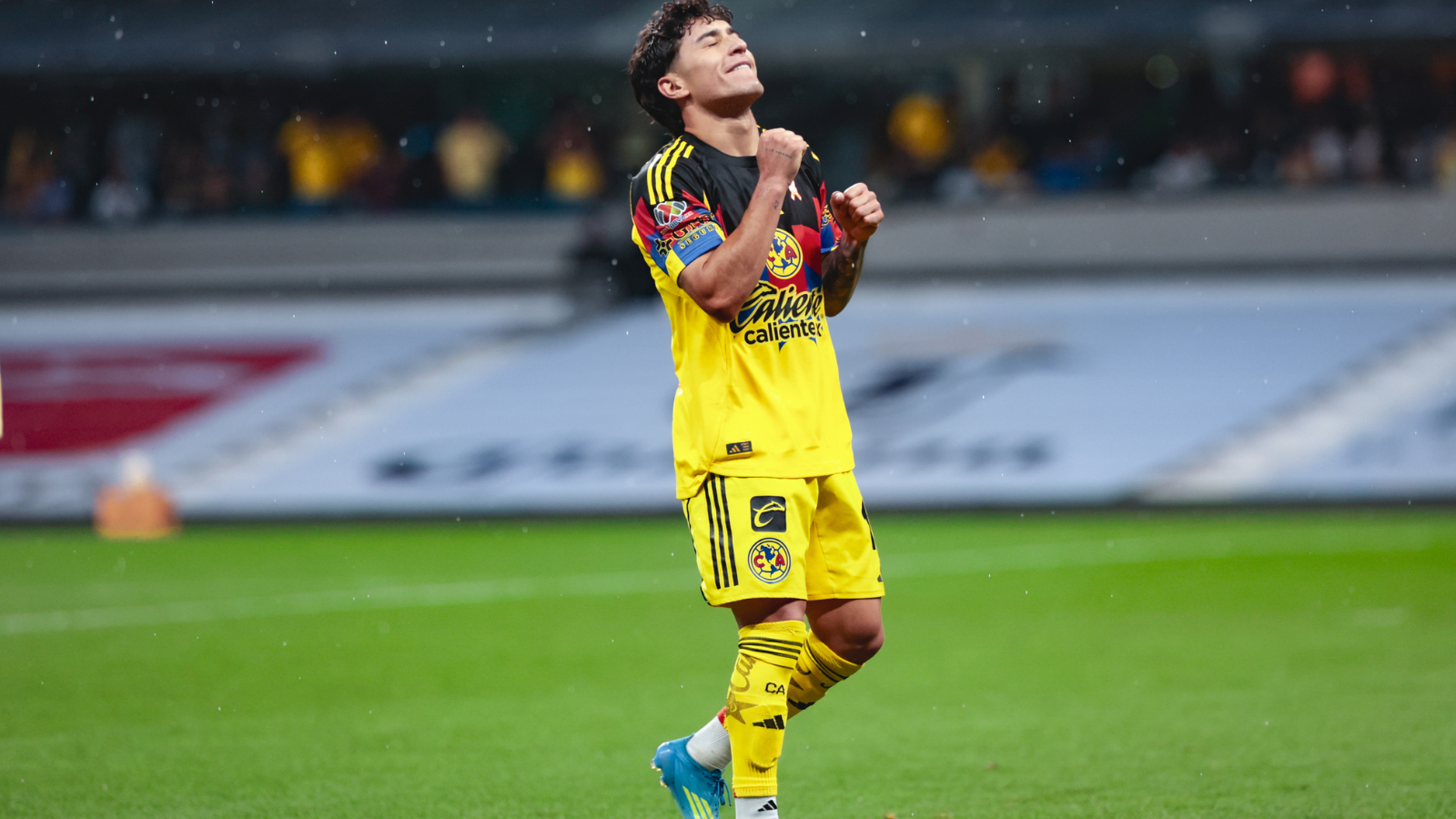 Soccer player in a yellow kit celebrating with clenched fists on a rain-soaked pitch.