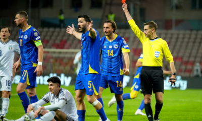 Soccer referee in yellow raises a red card as blue‑and‑yellow team players react on the field near an Italian team in white uniforms in a stadium.
