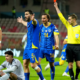 Soccer referee in yellow raises a red card as blue‑and‑yellow team players react on the field near an Italian team in white uniforms in a stadium.