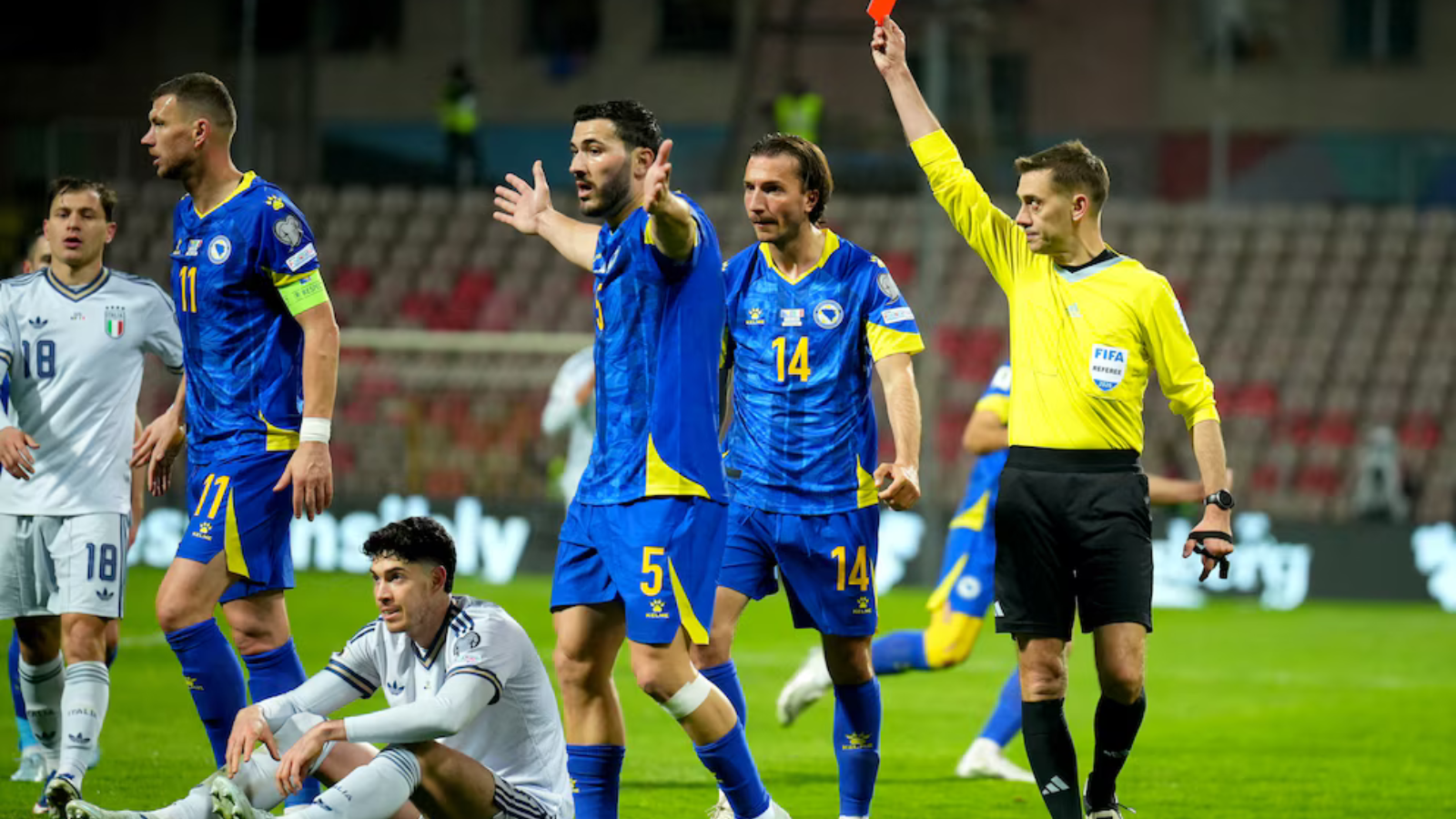 Soccer referee in yellow raises a red card as blue‑and‑yellow team players react on the field near an Italian team in white uniforms in a stadium.