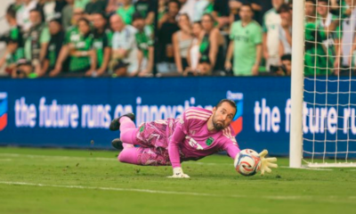 Soccer goalkeeper in a pink kit dives to his left to save the ball near the goal, with spectators in the background.