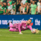 Soccer goalkeeper in a pink kit dives to his left to save the ball near the goal, with spectators in the background.