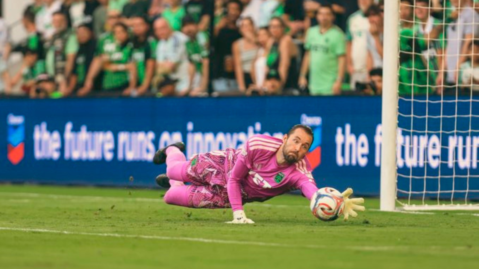Soccer goalkeeper in a pink kit dives to his left to save the ball near the goal, with spectators in the background.