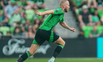 Soccer player in a green and black kit sprinting on the field during a match, blurred crowd in the stands behind.