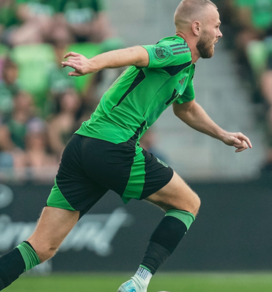 Soccer player in a green and black kit sprinting on the field during a match, blurred crowd in the stands behind.