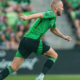 Soccer player in a green and black kit sprinting on the field during a match, blurred crowd in the stands behind.